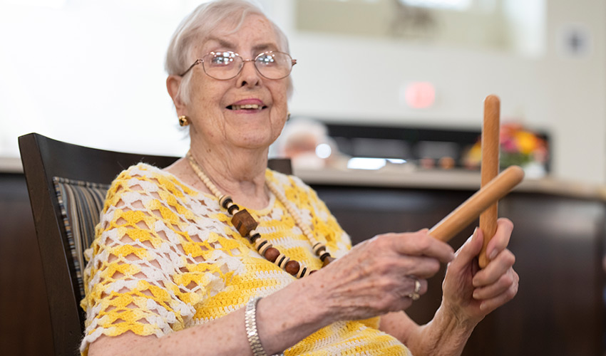 A senior woman sitting in a chair hitting two sticks together.