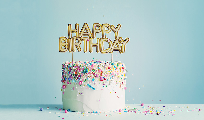 A white birthday cake with sprinkles and a sign on top.