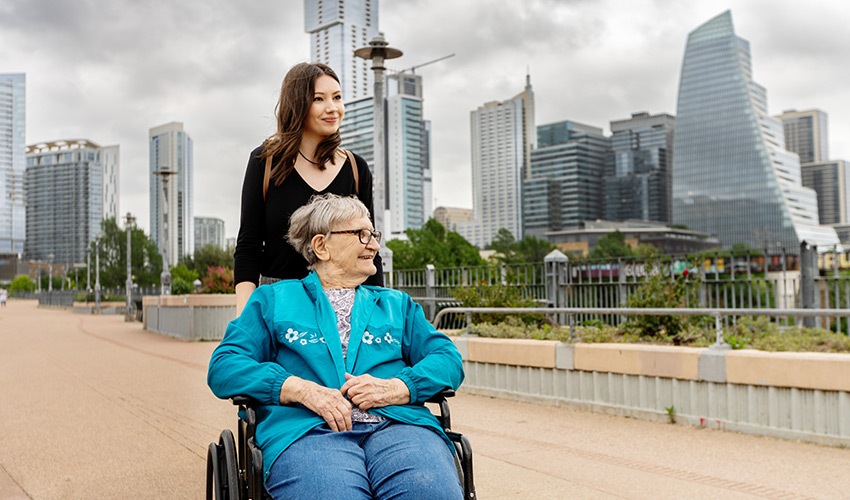 A person pushing another in a wheelchair with a city in the background.