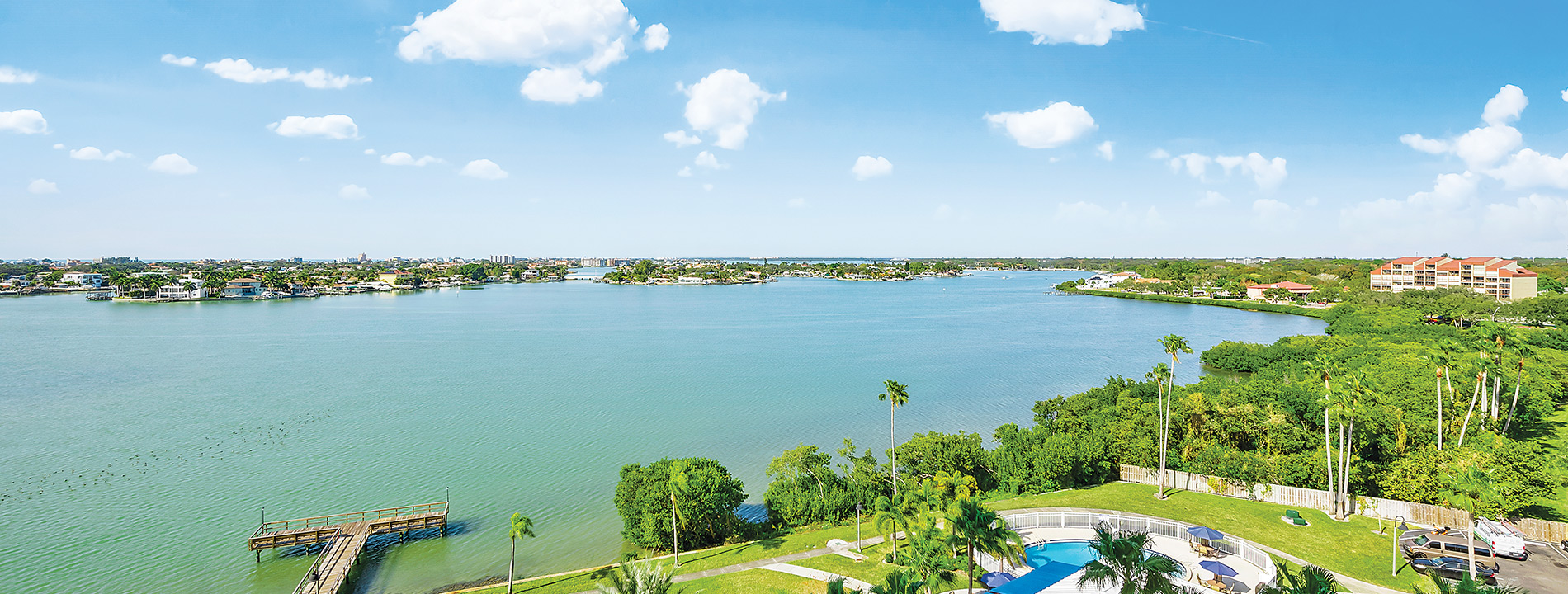 An aerial view of the property along the waters edge with palm trees and the city in the distance.