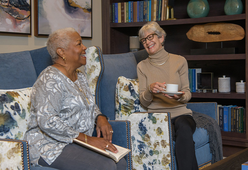 two residents are sitting in chairs in a common area laughing, holding a book and drinking tea