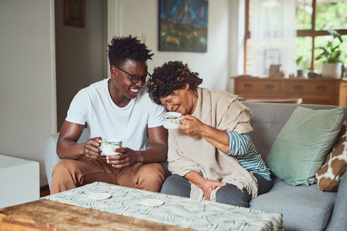 A young man and his elderly mother are sitting on a couch while holding their tea cups and laughing to one another.