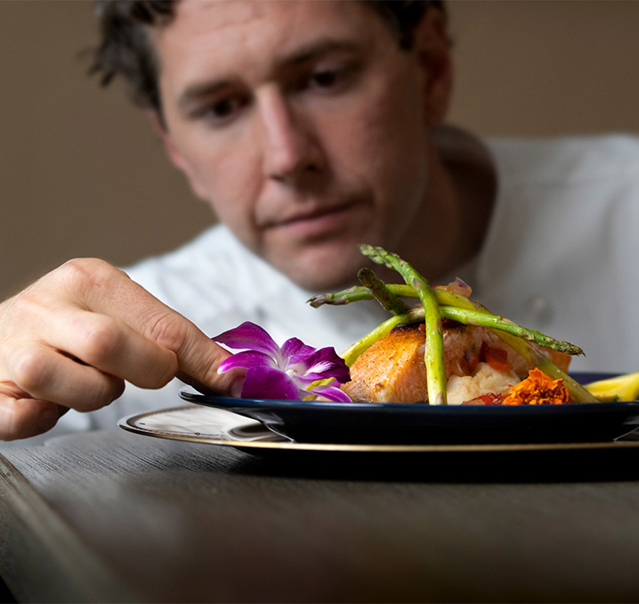 A chef placing a decorative flower on the edge of a meal to enhance the presentation A chef placing a decorative flower on the edge of a meal to enhance the presentation.