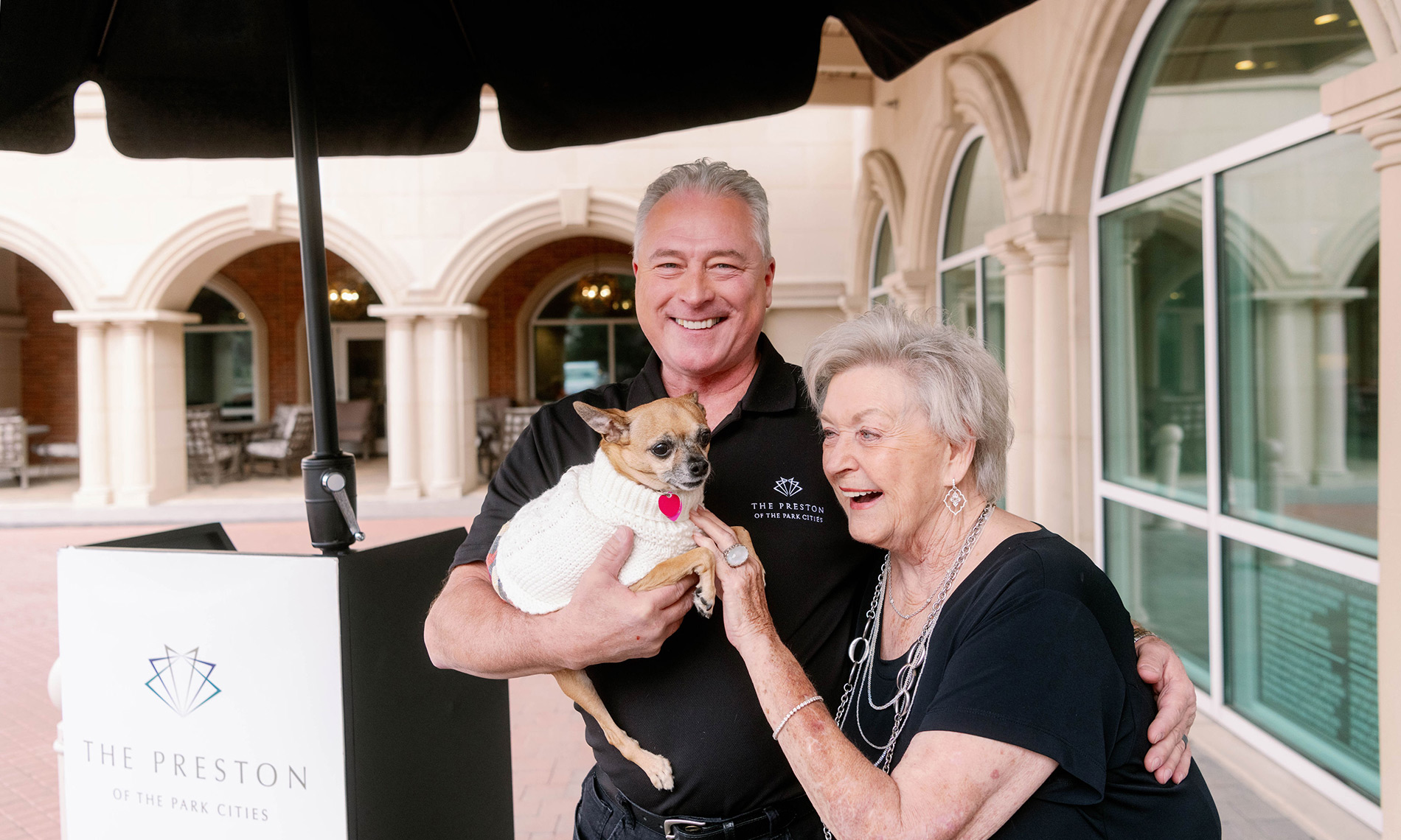 a smiling couple with their dog that is wearing a sweater sitting on the patio area