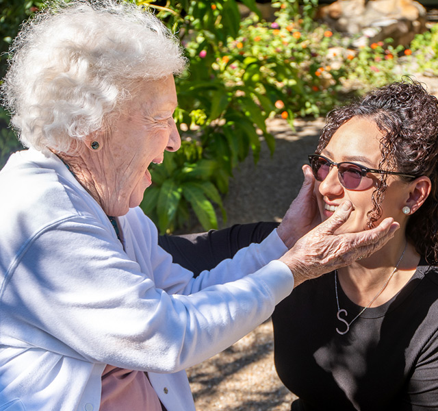 A resident outside is placing her hands on another resident's face smiling and laughing