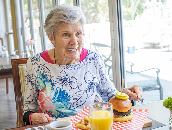 A resident is sitting at a dining table enjoying a burger and glass of orange juice.