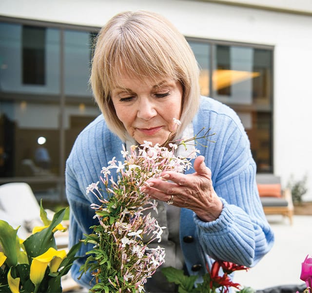 A resident in a blue sweater leaning over to smell light pink flowers in the garden