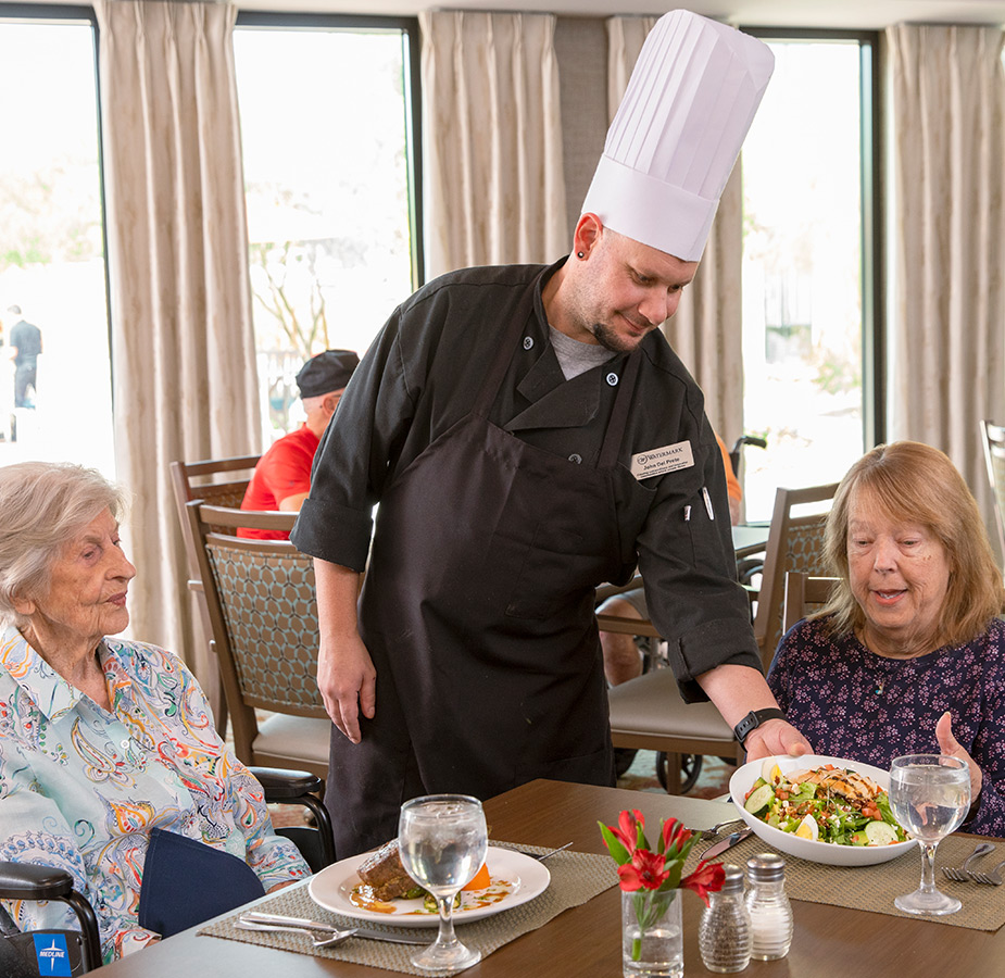 A chef is placing a plate of food down in front of a resident.